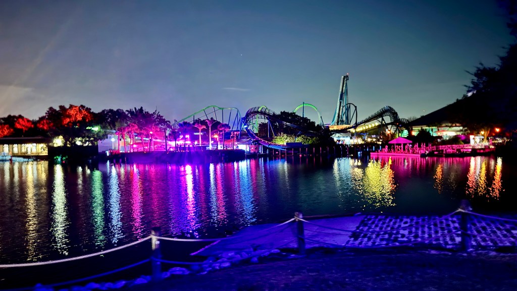 Parque de diversões noturno com montanhas-russas iluminadas em verde e azul, refletindo na água escura. Árvores e construções à esquerda e centro brilham em tons de vermelho, roxo e rosa, criando um cenário vibrante