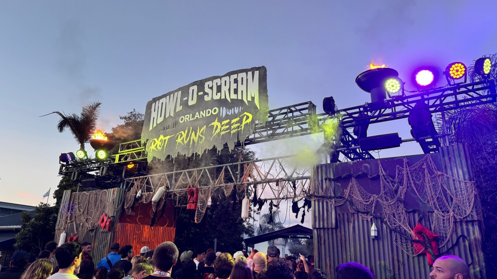 Entrada do evento Howl-O-Scream Orlando, com letreiro grande e iluminado, tochas acesas, fumaça verde e luzes coloridas. A estrutura é decorada com redes de pesca e boias vermelhas, e uma multidão de pessoas aguarda para entrar