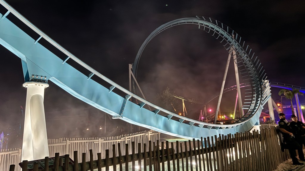 Montanha-russa azul-clara com looping e trilhos em espiral, iluminada por luzes coloridas e névoa no parque noturno. Dois policiais de uniforme escuro observam a cena à direita, perto de uma cerca de madeira e palmeiras. O céu está escuro