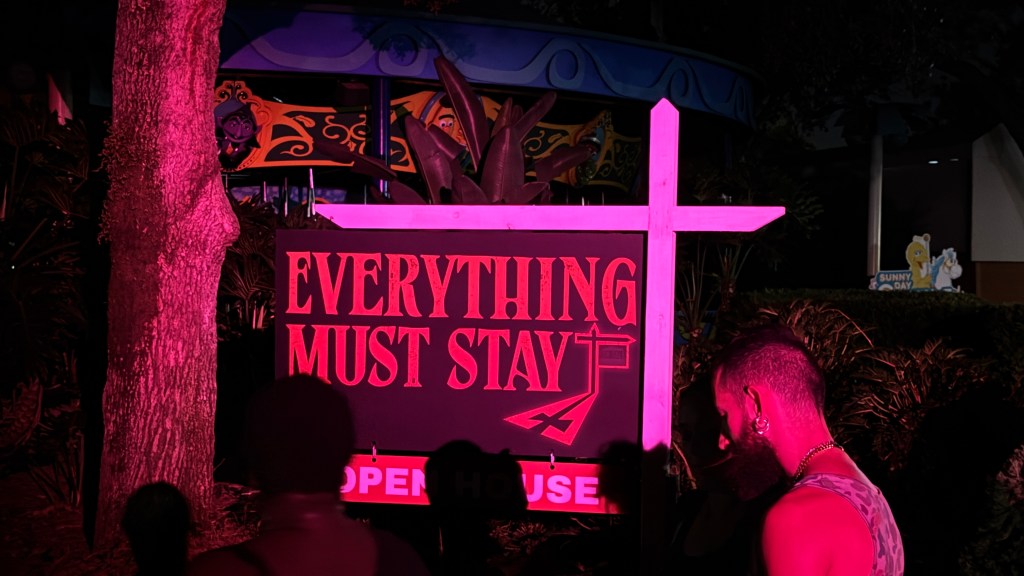 Placa preta com a frase EVERYTHING MUST STAY em letras vermelhas, iluminada por uma luz rosa forte. Uma cruz rosa neon está à direita da placa. Pessoas com silhuetas escuras e um tronco de árvore rosa estão em primeiro plano. Ao fundo, um carrossel e um letreiro SUNNY DAY com personagens de desenho animado são visíveis