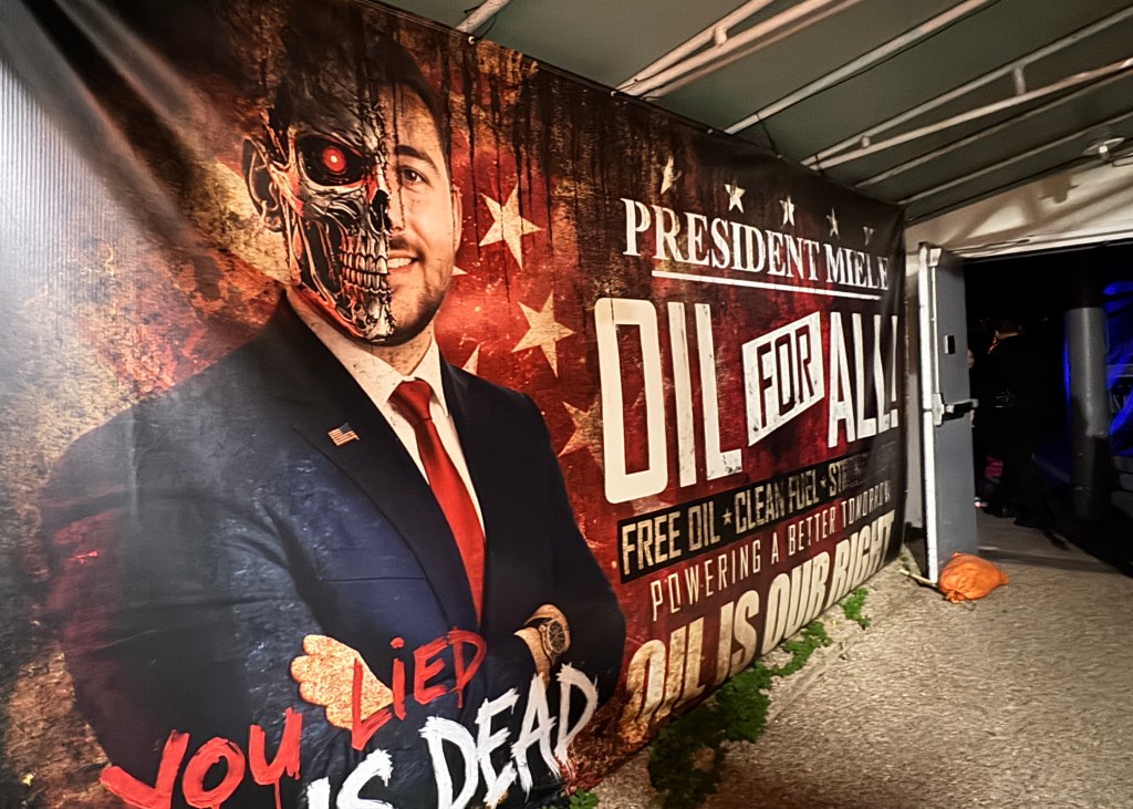 Banner de campanha política com um homem sorridente de terno azul e gravata vermelha, metade do rosto como um crânio robótico com olho vermelho. O fundo é uma bandeira americana desgastada. Textos: PRESIDENT MIELE, OIL FOR ALL!, FREE OIL + CLEAN FUEL = STRENGTH, POWERING A BETTER TOMORROW, OIL IS OUR RIGHT e YOU LIED IS DEAD