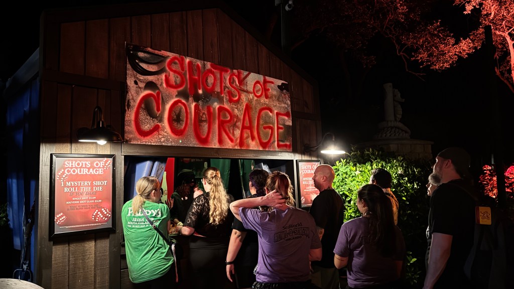Pessoas em fila noturna para um quiosque de madeira com letreiro SHOTS OF COURAGE em vermelho grafite. Um cartaz ao lado oferece Mystery Shot e Roll the Die. Uma árvore iluminada em vermelho e uma estátua de cavalo-marinho ao fundo completam a cena