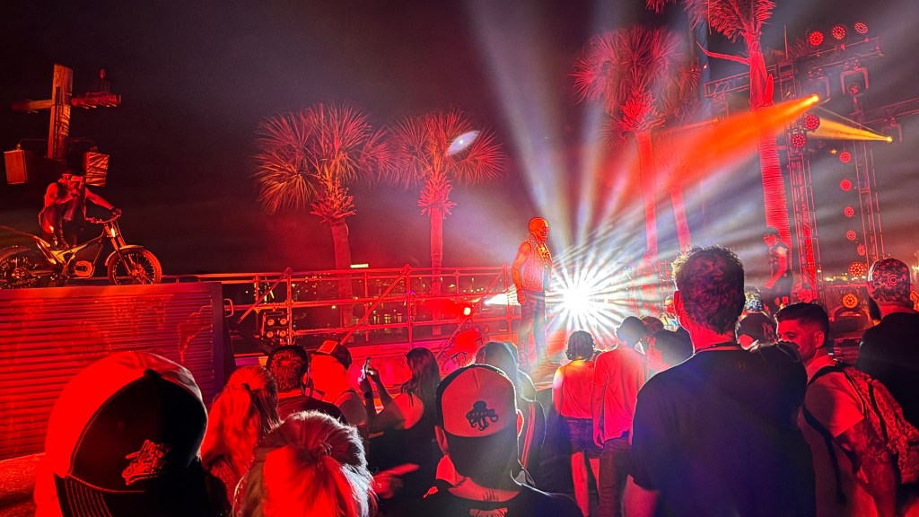 Pessoas assistem a um show noturno com luzes vermelhas intensas. Um artista de colete e calça escura está no palco, e outro, com capacete e mochila, está em uma moto ao lado de uma cruz de madeira. Palmeiras iluminadas e estruturas metálicas compõem o cenário, enquanto a plateia observa de costas, alguns com bonés e mochilas