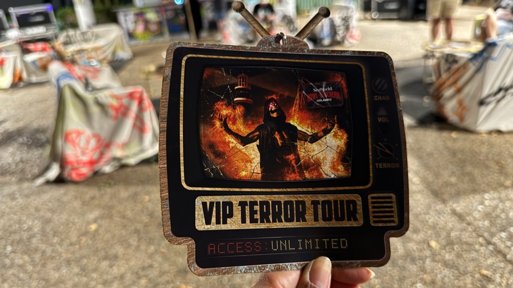 Mão segura um ingresso em formato de TV retrô com a imagem de uma figura sombria em chamas e os dizeres VIP TERROR TOUR e ACCESS: UNLIMITED