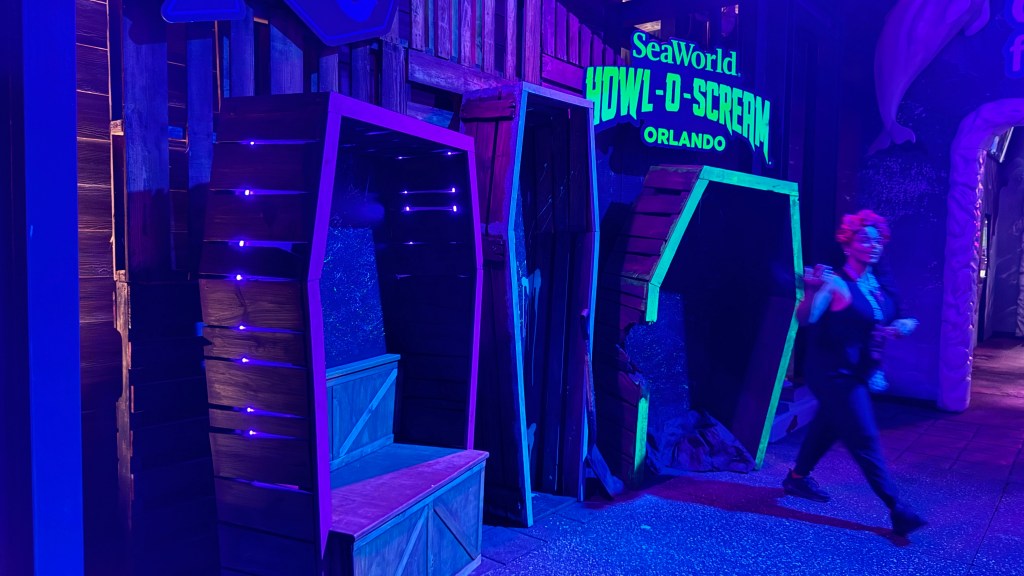 Cenário de Halloween com luzes neon roxas e azuis, caixões de madeira abertos e uma pessoa fantasiada de vampira com cabelo vermelho, maquiagem escura e roupa preta, caminhando à direita. Ao fundo, letreiro SeaWorld Howl-O-Scream Orlando em verde neon