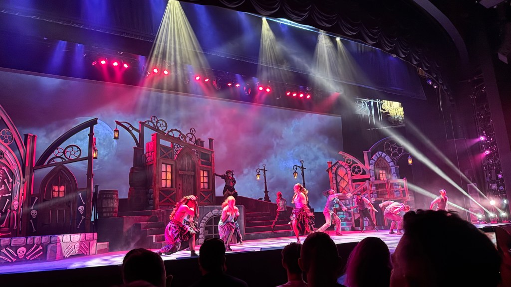 Palco teatral com cenário gótico de casas e esqueletos, iluminado por luzes vermelhas e azuis. Vários dançarinos, alguns com roupas escuras e outros com tons claros, executam movimentos coreografados. Uma pessoa em destaque no centro superior do palco aponta para a direita. O público assiste em primeiro plano