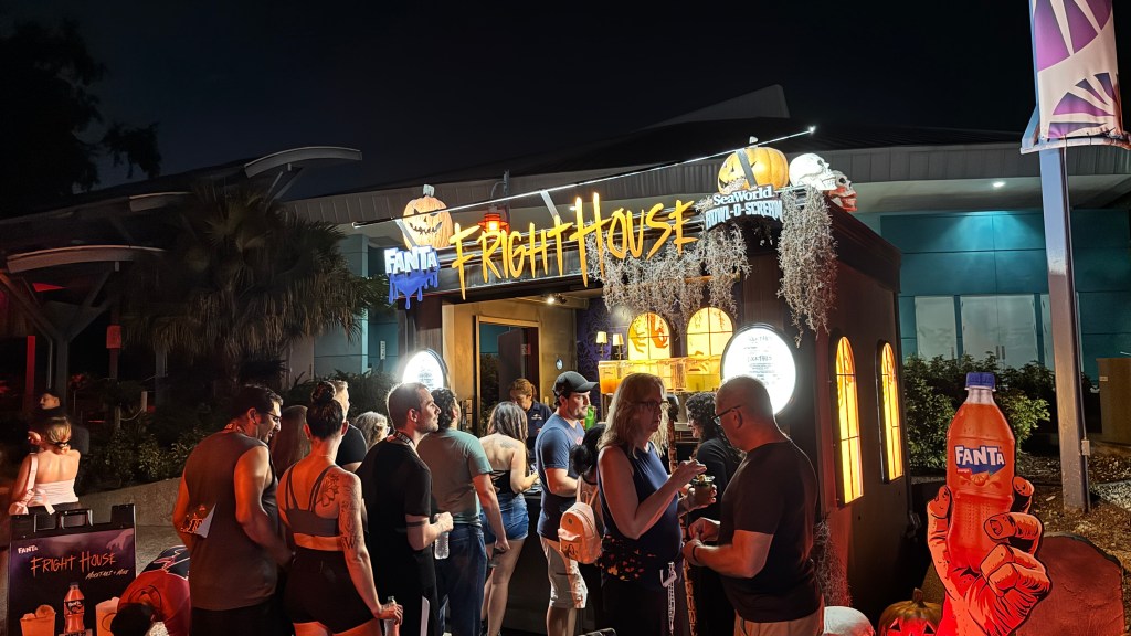 Pessoas em frente à Fright House do SeaWorld Howl-O-Scream, com letreiro iluminado, abóboras e caveiras decorativas. Uma mão gigante de Halloween segura uma garrafa de Fanta laranja.