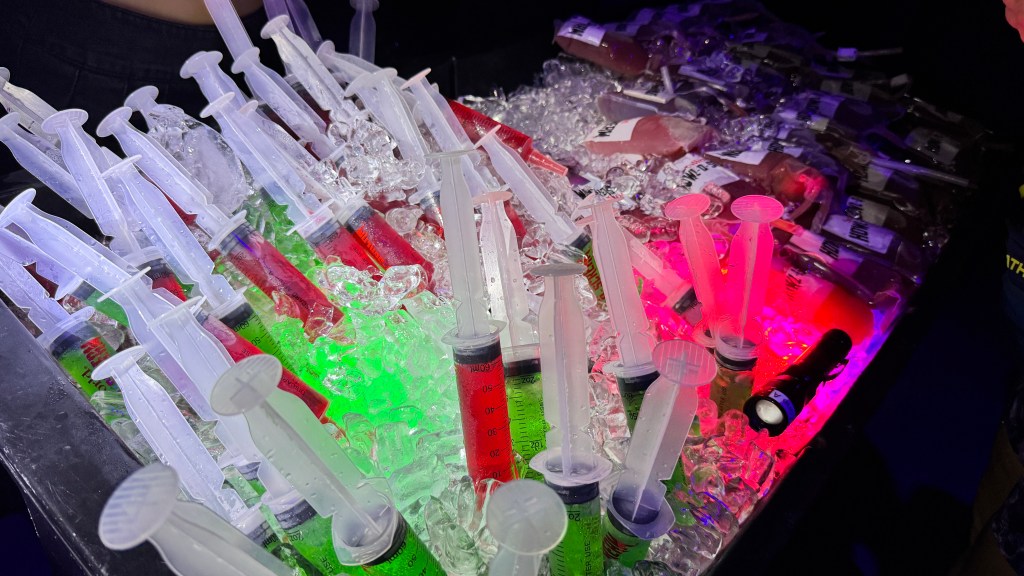 Seringas plásticas transparentes cheias de líquidos coloridos, vermelho e verde, imersas em gelo picado, iluminadas por luzes neon vermelhas e verdes. Ao fundo, garrafas de bebidas também em gelo, com uma lanterna preta acesa. O ambiente é escuro, sugerindo uma festa ou evento noturno