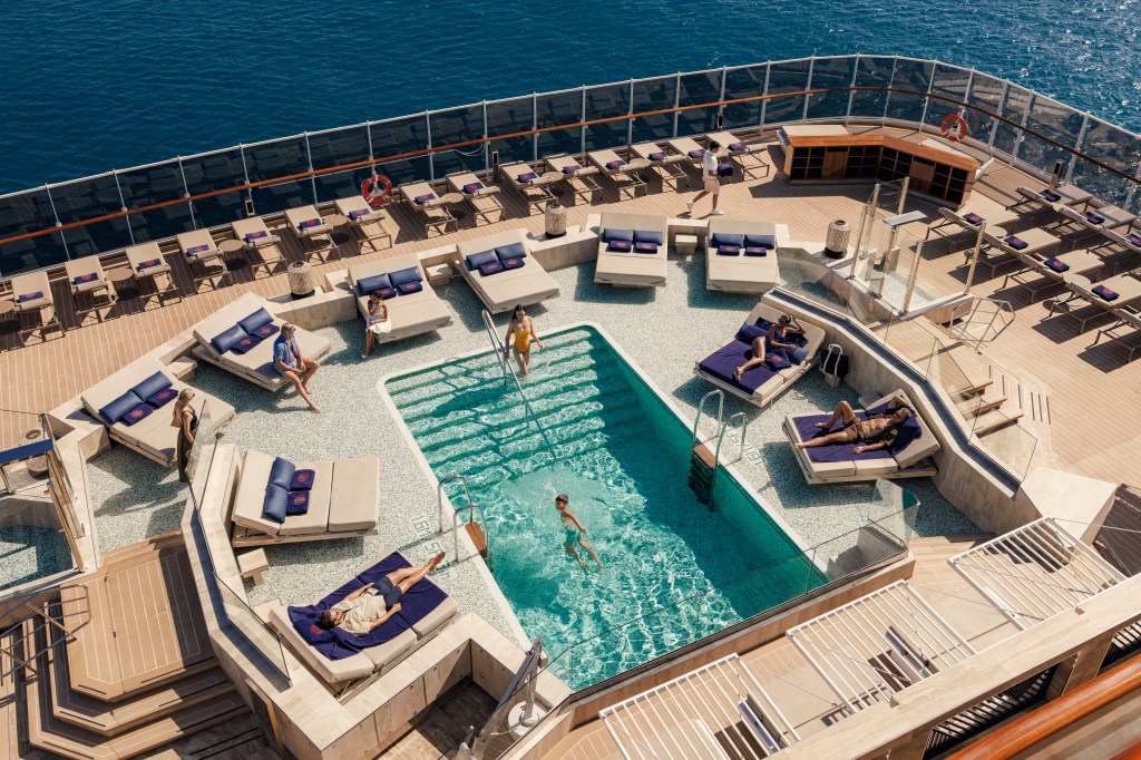 Vista aérea de um deck de navio com piscina retangular de água azul-turquesa, cercada por espreguiçadeiras e camas de solteiro. Pessoas relaxam, nadam e caminham pelo deck de madeira, sob um céu claro e mar azul ao fundo