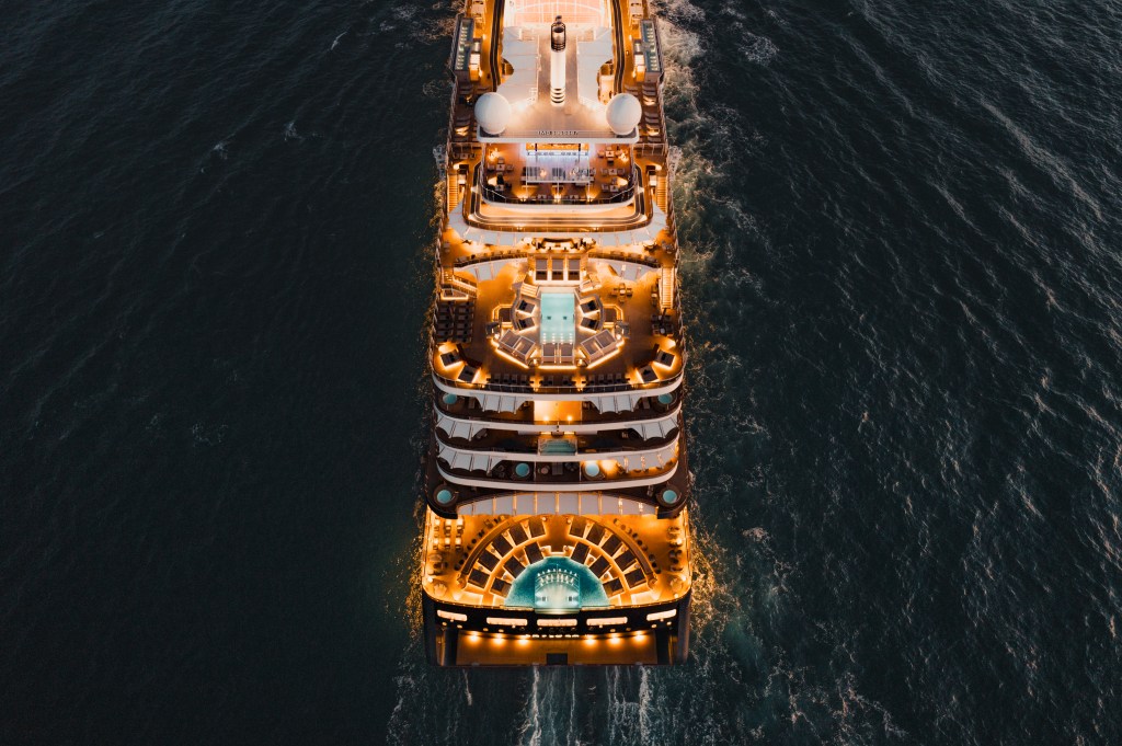 Navio de cruzeiro luxuoso, visto de cima, navegando em águas escuras à noite. As luzes quentes do navio iluminam as piscinas e decks, criando um rastro de espuma na água