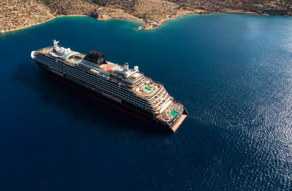 Navio de cruzeiro grande, preto e branco, com piscinas e espreguiçadeiras no convés, navegando em águas azuis profundas perto de uma costa rochosa com vegetação esparsa e algumas casas brancas