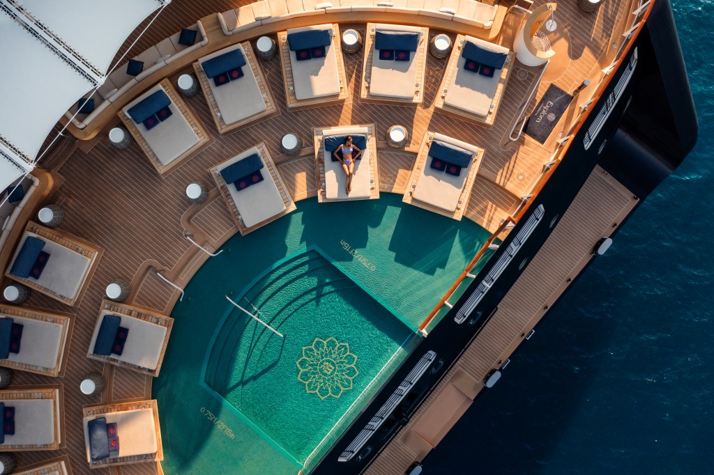 Vista aérea do convés de um navio de luxo com uma piscina azul-esverdeada em formato de meia-lua. Uma mulher de biquíni roxo está deitada em uma das espreguiçadeiras ao redor da piscina. Há várias espreguiçadeiras com almofadas claras e travesseiros azuis, além de mesas de apoio brancas. O convés é de madeira clara, e a água do mar azul-escuro cerca o navio