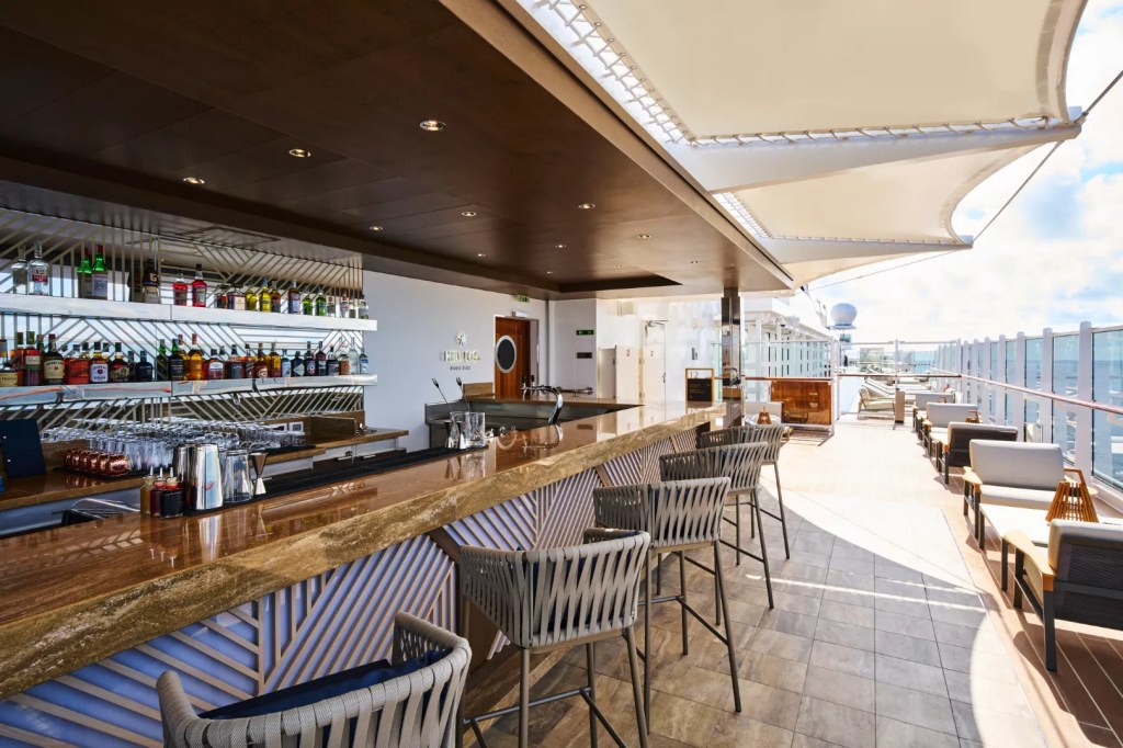 Bar de luxo ao ar livre em um navio, com balcão de madeira e base geométrica iluminada, banquetas estofadas e prateleiras com garrafas de bebidas. À direita, área de lounge com sofás e mesas, sob um toldo retrátil, com vista para o mar e céu azul