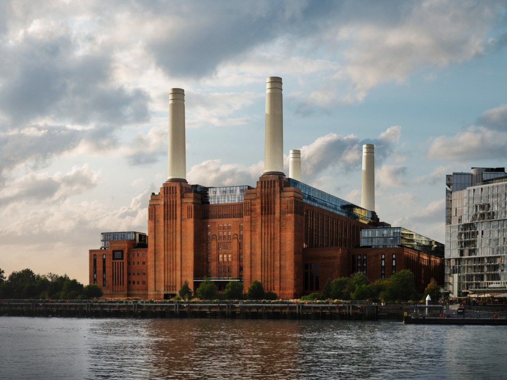 A Usina Termelétrica de Battersea, um grande edifício de tijolos vermelhos com quatro chaminés brancas, vista da água sob um céu parcialmente nublado