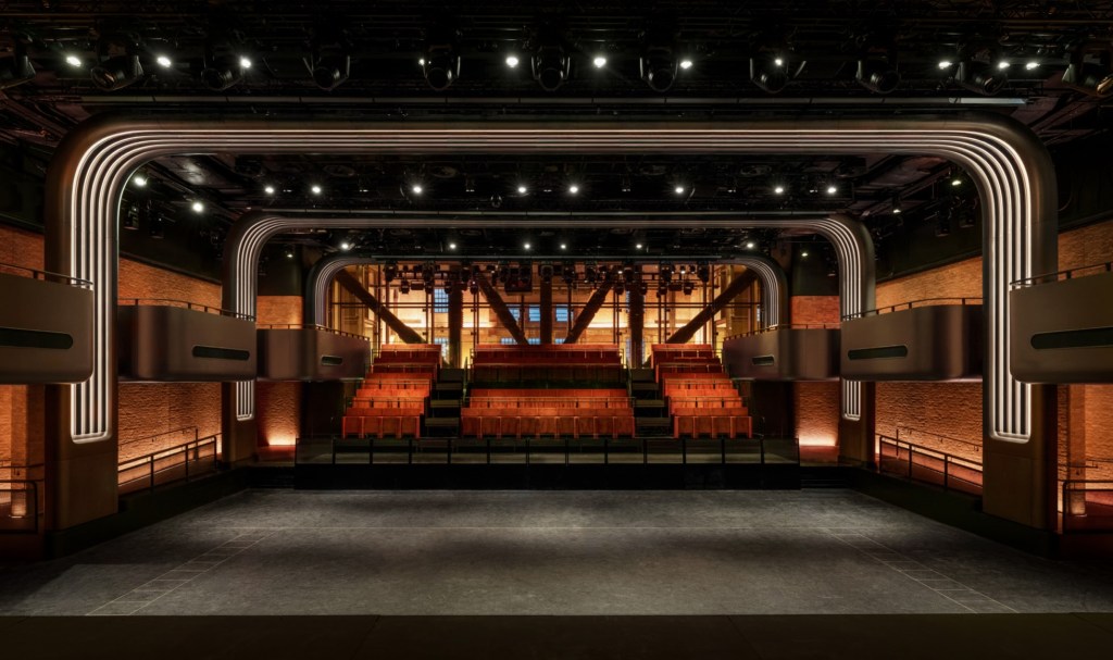 Interior de um teatro moderno com palco vazio em primeiro plano. Ao fundo, assentos em níveis ascendentes com estofamento laranja. Laterais com paredes de tijolos iluminadas em tom quente e camarotes suspensos com iluminação vertical embutida. O teto escuro possui diversos refletores focados no palco