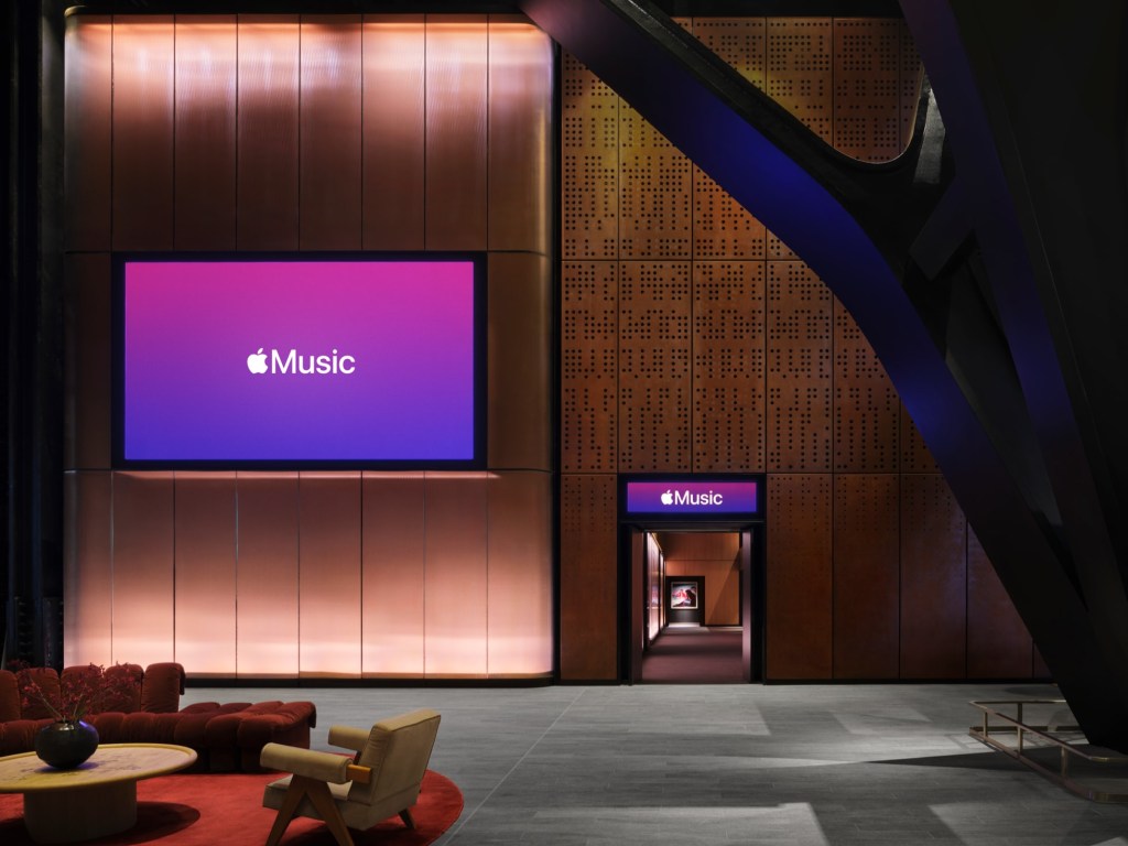 Interior moderno com tela grande exibindo o logo Apple Music em fundo roxo e rosa. Abaixo, sofás vermelhos e poltrona bege sobre tapete vermelho. À direita, parede texturizada e estrutura metálica escura, com uma porta iluminada também com o logo Apple Music levando a um corredor