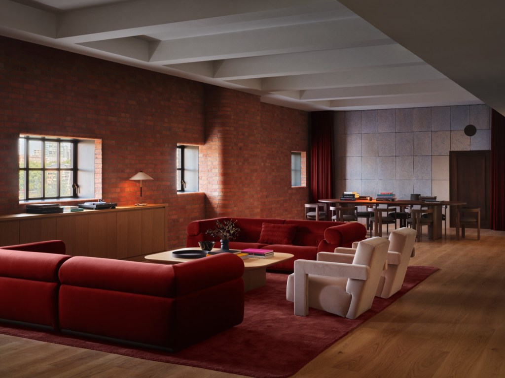 Sala de estar e jantar com paredes de tijolos aparentes, sofás vermelhos e duas poltronas claras sobre um tapete vinho. Ao fundo, uma mesa de jantar de madeira com cadeiras, cortinas vermelhas e parede cinza. Luz natural entra pelas janelas, iluminando o ambiente aconchegante