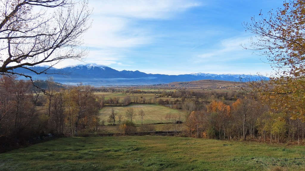 Paisagem outonal com campos verdes e marrons, árvores com folhagem amarelada e galhos secos. Ao fundo, montanhas com picos nevados sob um céu azul claro com nuvens esparsas.