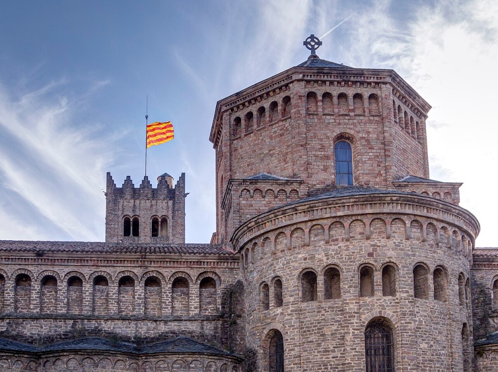 Vista parcial de uma construção medieval de pedra, com uma torre ameada à esquerda exibindo uma bandeira catalã de listras vermelhas e amarelas, e um edifício octogonal à direita com uma cruz celta no topo, sob um céu azul com nuvens esparsas