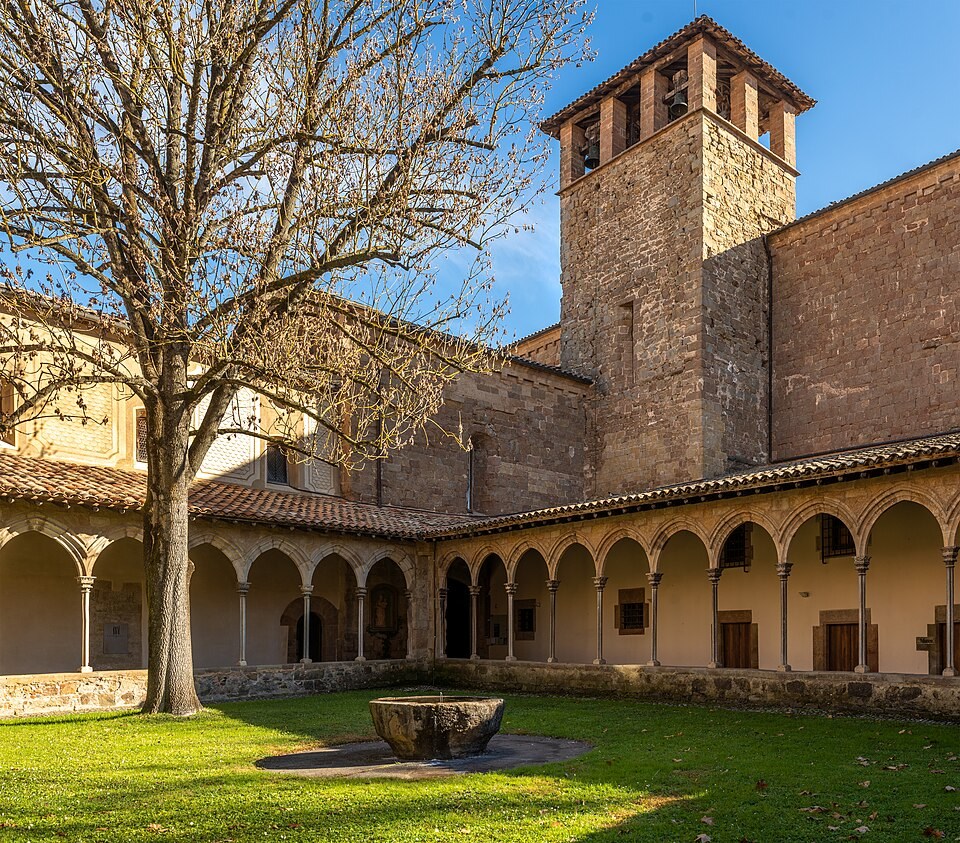 Pátio interno de um mosteiro com gramado verde, uma árvore sem folhas no centro e uma fonte de pedra. Ao redor, um claustro com arcos de pedra e telhado de telhas marrons, e ao fundo, uma torre sineira de pedra sob céu azul