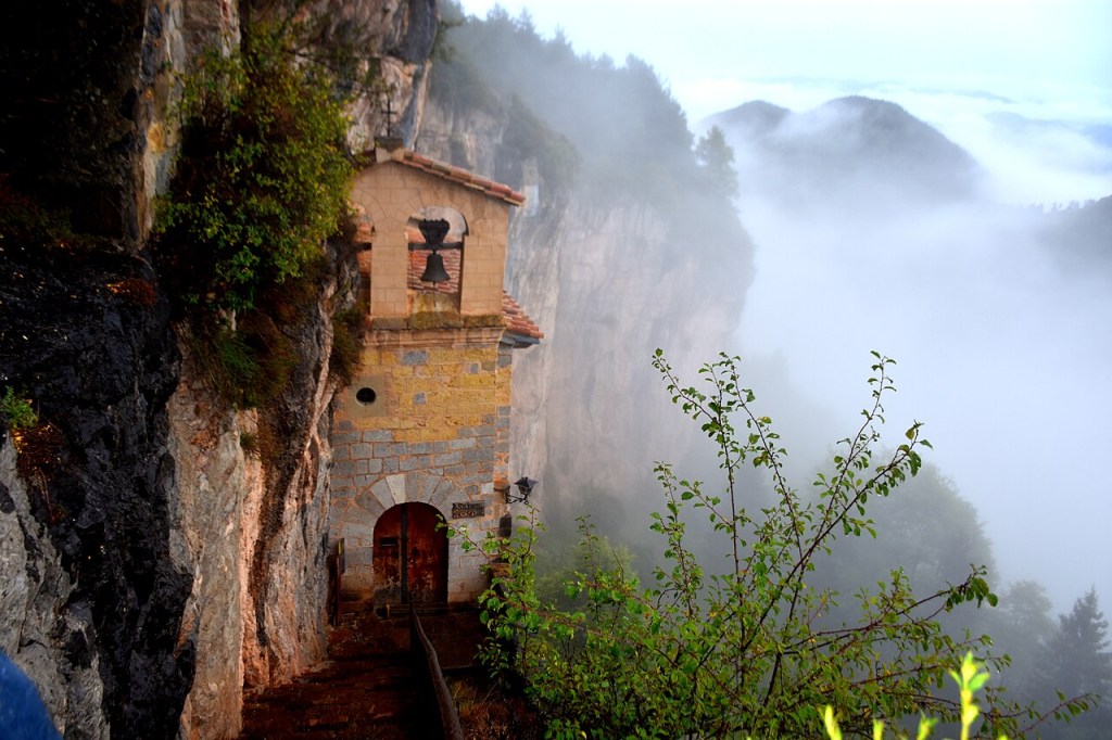 Pequena capela de pedra com telhado de telhas e um sino, construída na lateral de um penhasco rochoso. Uma porta de madeira escura e um caminho estreito levam à entrada. À direita, a paisagem montanhosa está coberta por uma névoa densa, com árvores escuras visíveis ao fundo. Folhagens verdes em primeiro plano.