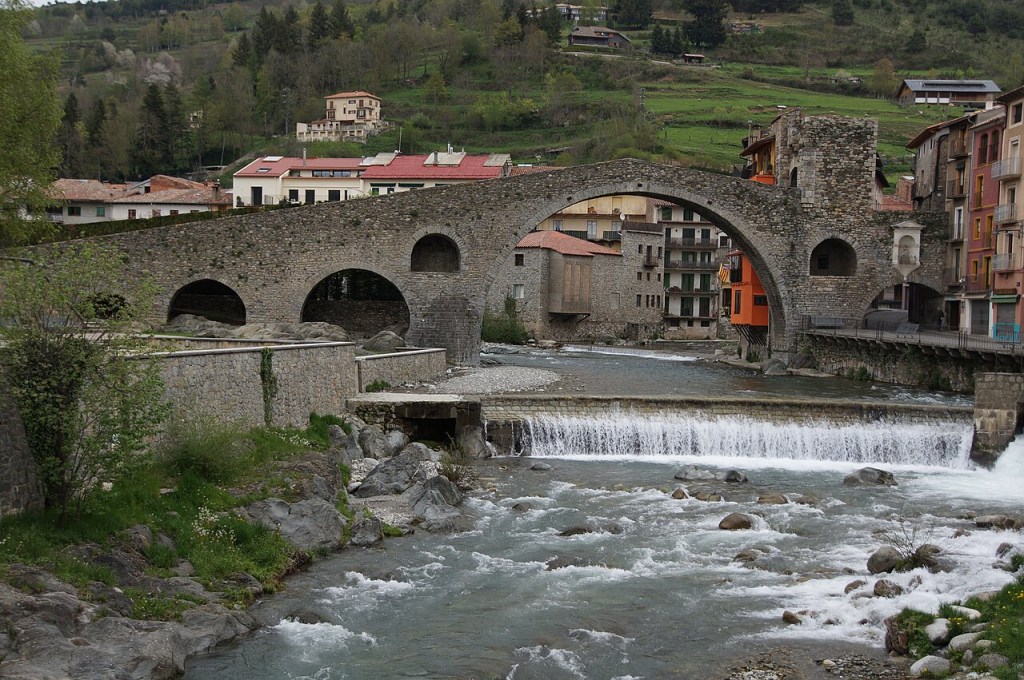 Ponte medieval de pedra com arcos sobre um rio caudaloso, com uma pequena cachoeira. Casas coloridas e montanhas verdes ao fundo, sob céu nublado