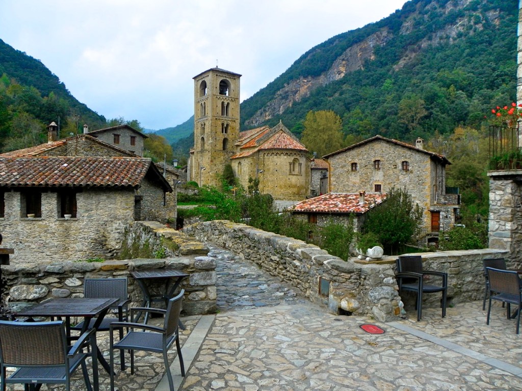 Vila medieval com casas de pedra e telhados de cerâmica, uma torre sineira alta e uma igreja românica ao fundo. Em primeiro plano, mesas e cadeiras de um café em um pátio de pedras, com uma ponte de pedra à esquerda e montanhas arborizadas sob um céu nublado