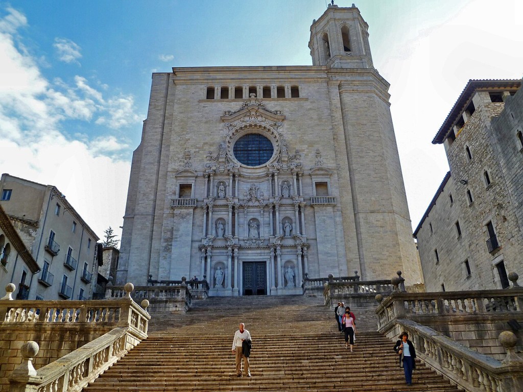 Escadaria de pedra com pessoas subindo e descendo, levando à fachada ornamentada da Catedral de Girona, com uma rosácea central e torre sineira, sob céu azul com nuvens esparsas
