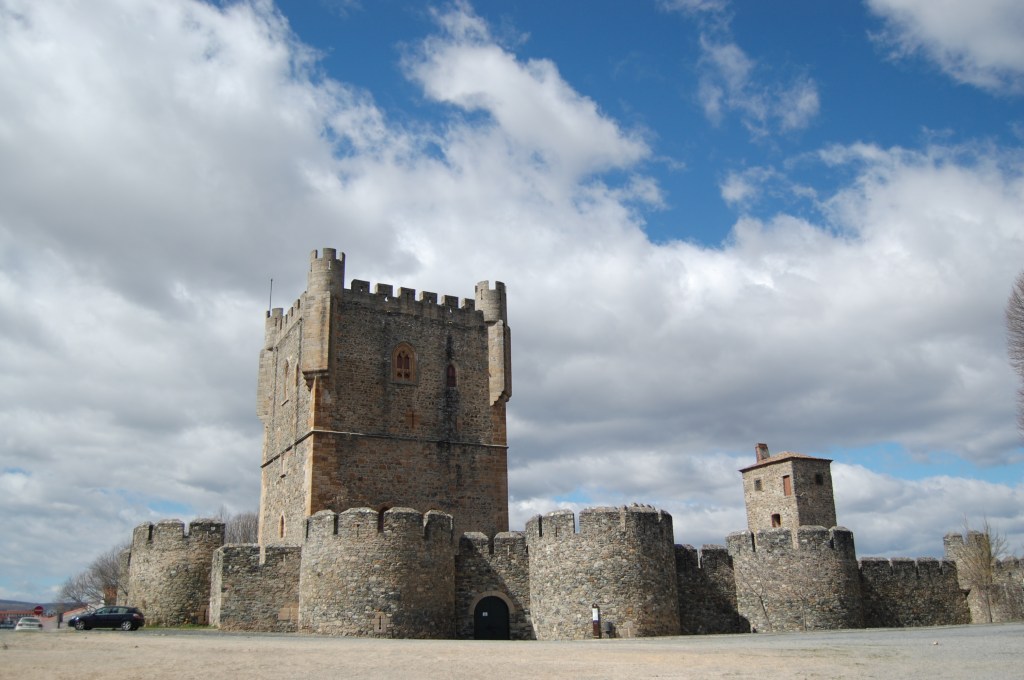 Castelo medieval de pedra com torre principal alta e ameias, muralhas robustas e uma torre menor à direita, sob céu parcialmente nublado com nuvens brancas e azuis. Um carro escuro está estacionado à esquerda, próximo à base da muralha.