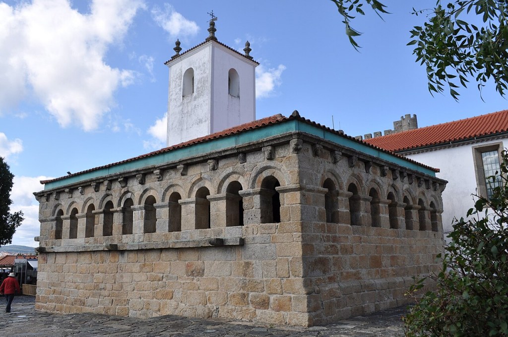 Igreja românica de pedra com arcos e telhado de telhas, torre sineira branca ao fundo, céu azul com nuvens e folhagem verde à direita