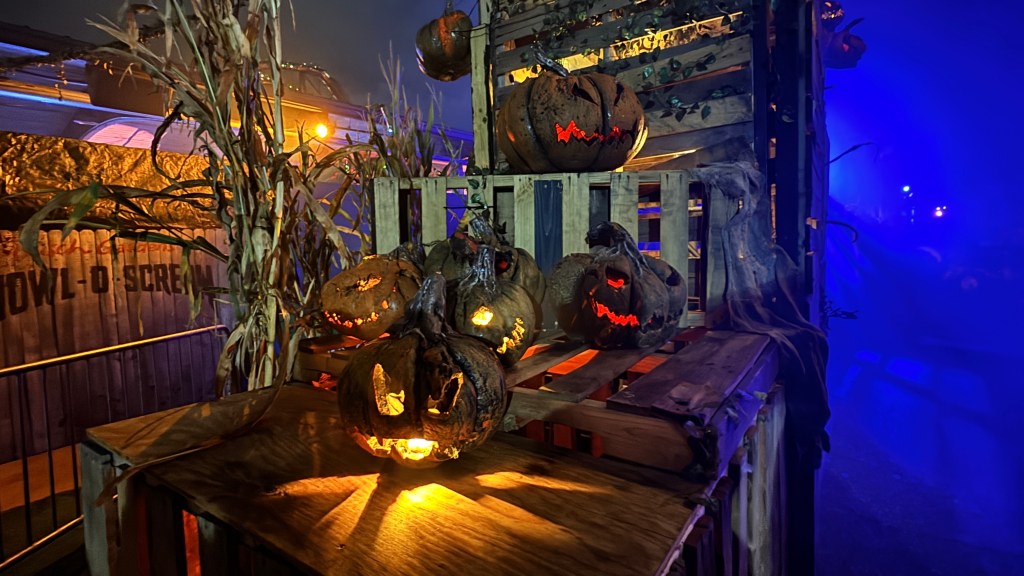Decoração de Halloween com abóboras escuras e iluminadas, algumas com rostos assustadores, sobre caixotes de madeira. Há plantas secas de milho e um letreiro HOWL-O-SCREAM ao fundo, com névoa azul à direita