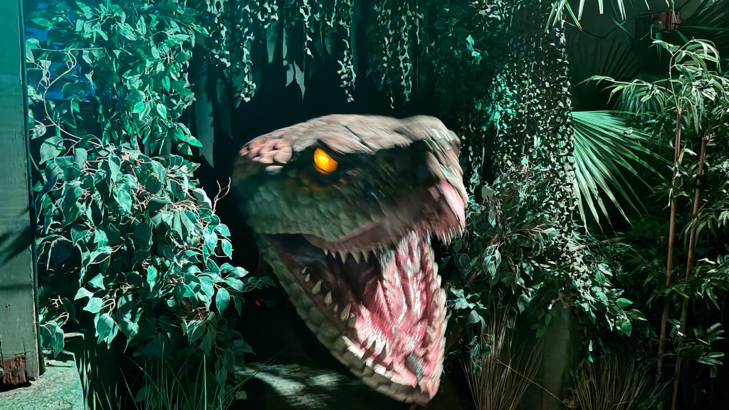 Cabeça de dinossauro animatrônico com olhos amarelos brilhantes e boca aberta, exibindo dentes afiados, emergindo de uma folhagem densa e escura, simulando uma selva noturna