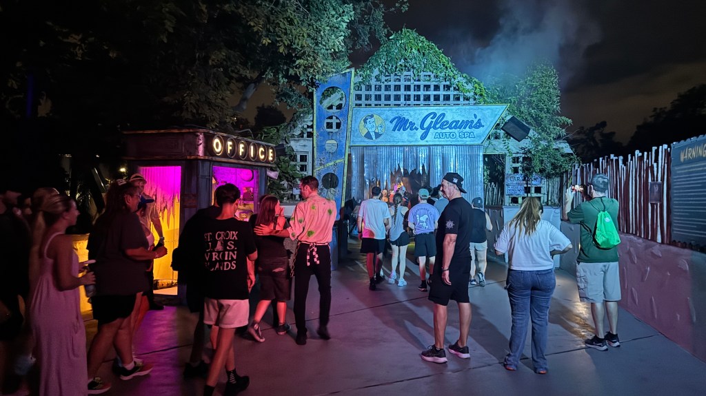 Pessoas em um evento noturno, com iluminação roxa e azul. À esquerda, um prédio com a palavra OFFICE iluminada em amarelo. Ao centro, uma fachada com Mr. Gleams Auto Spa e folhagens. À direita, uma cerca de madeira e um painel com WARNING. O ambiente é escuro, com fumaça no ar, criando uma atmosfera misteriosa