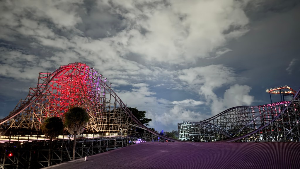 Montanha-russa de madeira iluminada em tons de vermelho e roxo à noite, com céu nublado e palmeiras na base