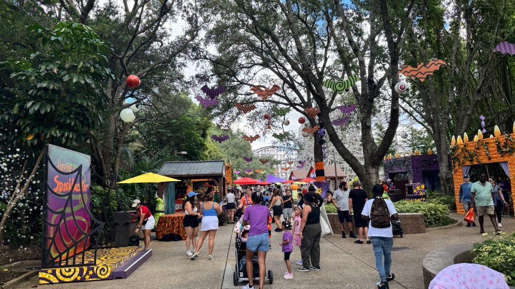 Parque temático decorado para o Halloween, com árvores enfeitadas com morcegos coloridos e olhos gigantes. Pessoas caminham por uma alameda com barracas de comida e bebida, e ao fundo, uma montanha-russa branca. Um palco com teia de aranha e a frase Having a Spooktacular Day aparece à esquerda