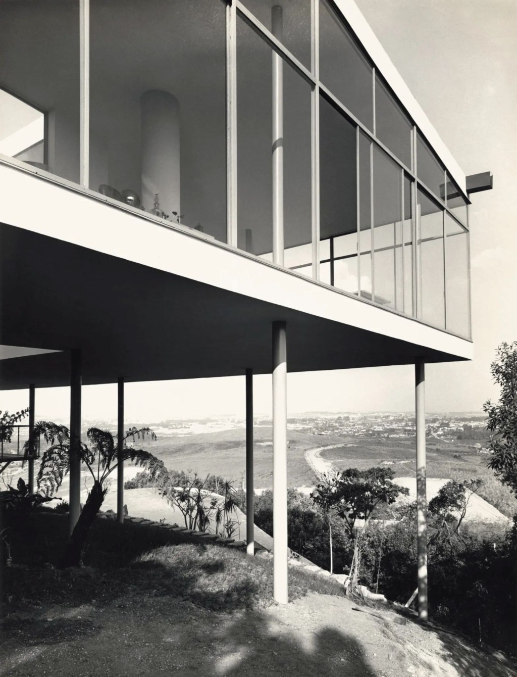 Casa modernista em preto e branco, suspensa por pilares finos, com fachada de vidro refletindo o céu. Abaixo, um gramado inclinado com árvores e, ao fundo, uma paisagem urbana com estradas sinuosas e vegetação.
