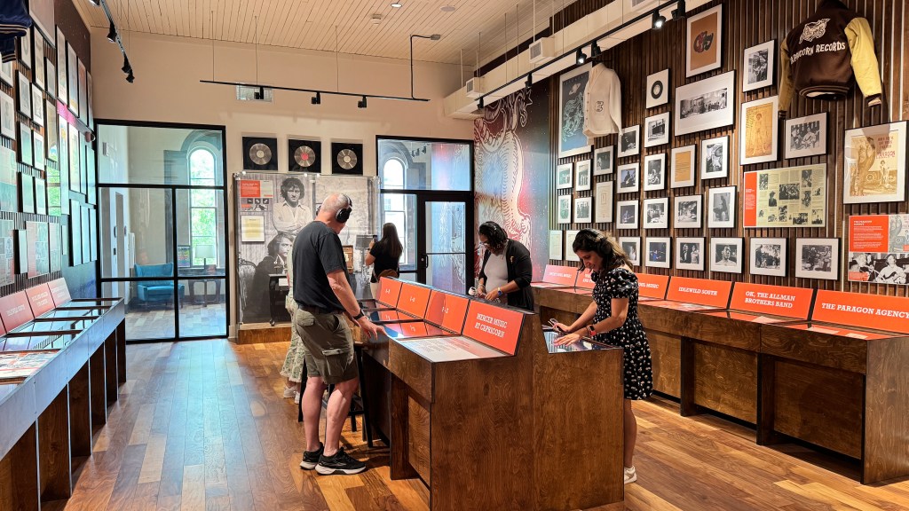 Pessoas visitam um museu de música com paredes repletas de quadros e discos de vinil. Um homem de camiseta preta e bermuda cáqui usa fones de ouvido e interage com uma tela interativa. Uma mulher de vestido estampado e fones de ouvido também interage com outra tela.
