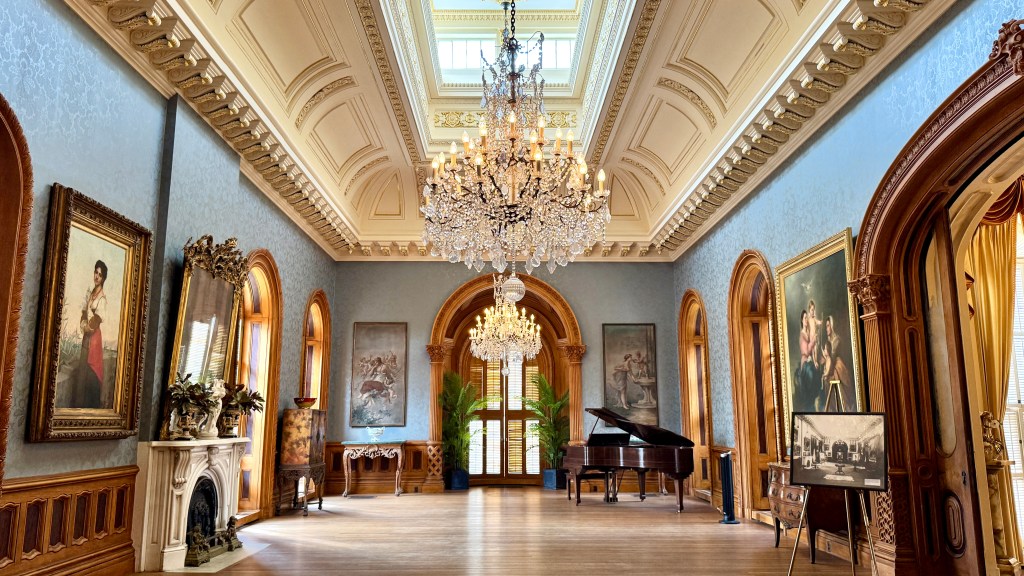 Salão luxuoso com paredes azuis e rodapés de madeira, teto ornamentado com lustres de cristal, janelas arqueadas e pinturas clássicas, incluindo um piano de cauda e uma lareira branca