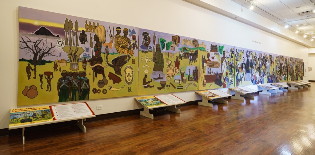 Mural colorido em uma galeria, retratando a história e cultura africana desde suas origens até os dias atuais, com figuras humanas, animais e objetos. Abaixo, expositores com informações complementares sobre a arte