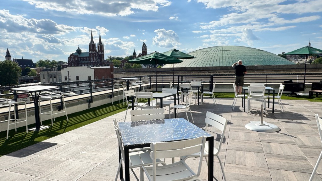 Terraço com mesas e cadeiras brancas, um homem de costas olhando a paisagem urbana com uma igreja gótica e um domo verde ao fundo, sob céu azul com nuvens