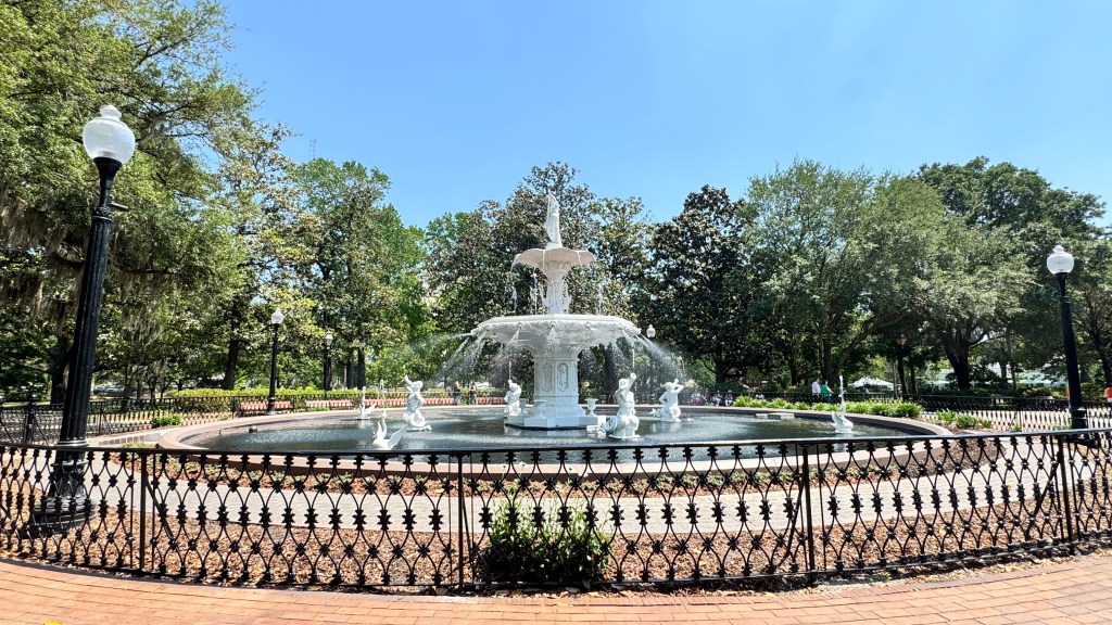 Fonte branca ornamentada com estátuas de figuras mitológicas jorrando água, cercada por um lago e um gradil de ferro preto. Ao fundo, árvores frondosas e um céu azul claro, com postes de luz antigos e pessoas caminhando no parque