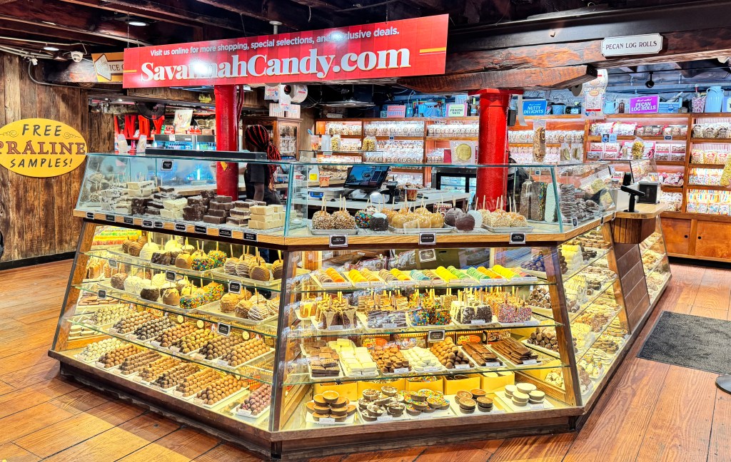 Interior de uma loja de doces, Savannah Candy, com um grande balcão de vidro exibindo diversos doces, como maçãs caramelizadas, trufas, bombons e fudge. Ao fundo, prateleiras de madeira com mais produtos embalados e um letreiro vermelho com SavannahCandy.com. Uma pessoa com cabelo trançado está atrás do balcão. O chão é de madeira.