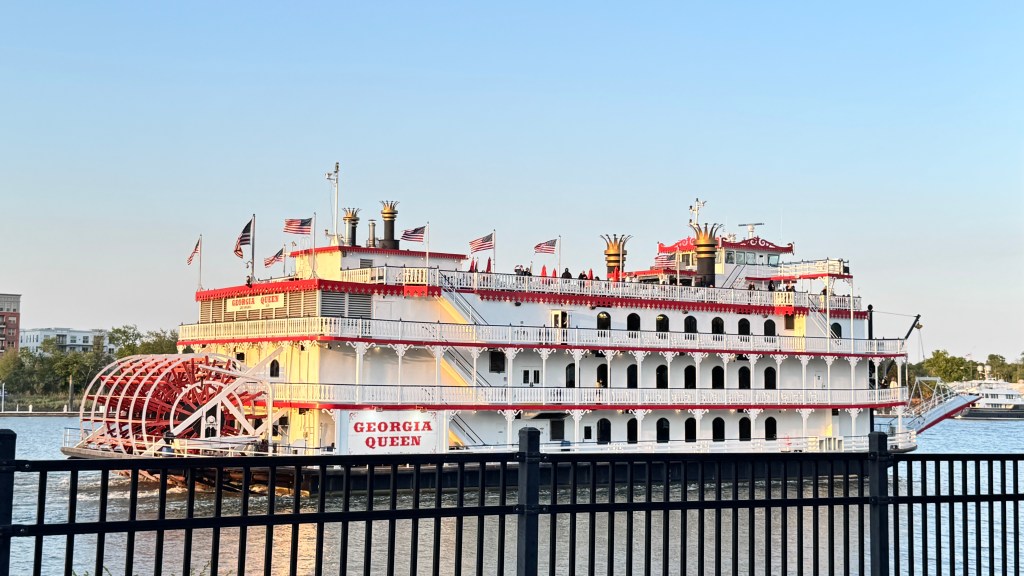 Barco a vapor Georgia Queen branco e vermelho, com uma grande roda dágua vermelha na popa, navegando em um rio sob céu azul claro. Bandeiras americanas tremulam nos mastros, e pessoas são visíveis nos decks superiores. Uma grade preta de metal está em primeiro plano, com edifícios ao fundo.
