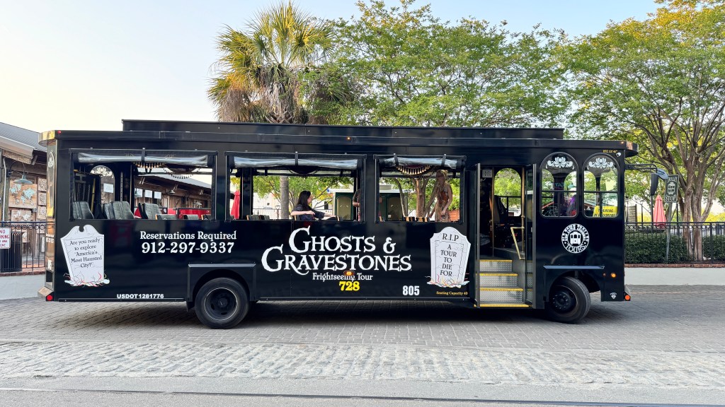 Ônibus turístico preto com Ghosts & Gravestones Frightseeing Tour em branco, número 728 em amarelo e 805 em branco. Duas mulheres sentadas, uma de blusa escura e outra de vestido estampado. O ônibus tem janelas abertas, assentos vazios e uma placa R.I.P. A Tour To Die For. Ao fundo, árvores e um céu claro. O veículo está em uma rua de paralelepípedos.