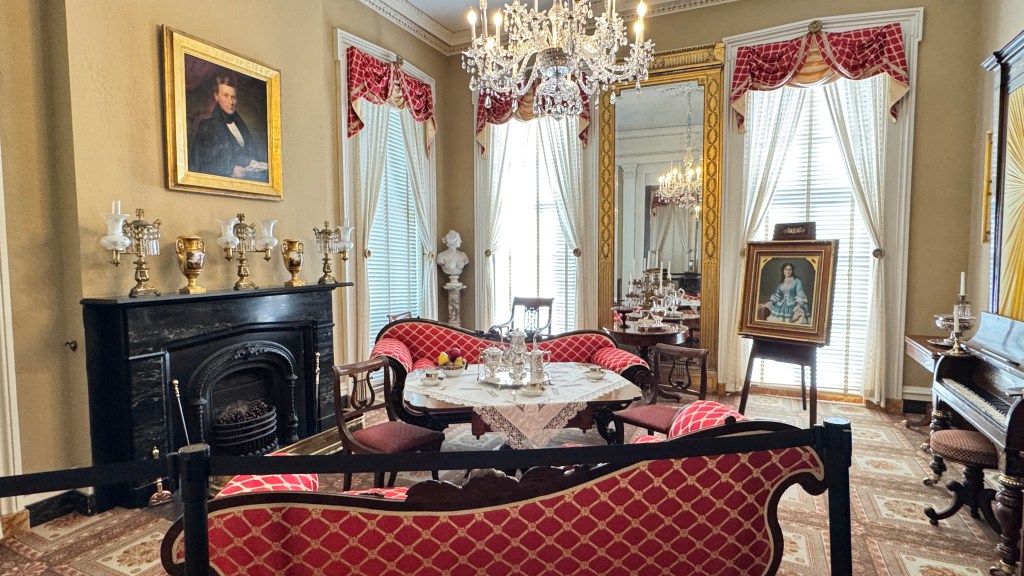 Sala de estar luxuosa com paredes bege, lareira preta, sofás vermelhos e mesa de centro com serviço de chá. Lustre de cristal, espelho grande, janelas com cortinas brancas e vermelhas, e quadros de época decoram o ambiente. Um piano escuro e uma estátua de busto branco completam a cena.