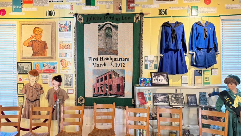 Exposição de uniformes e memorabilia das Girl Scouts, com um quadro de uma menina loira fazendo saudação, dois uniformes azuis de marinheiro e um manequim com uniforme verde, além de um tapete verde com a inscrição Juliette Gordon Low First Headquarters March 12, 1912 e imagens de edifícios