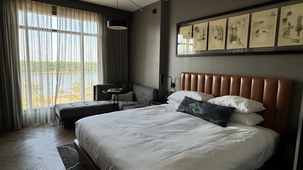Quarto de hotel com cama king-size de lençóis brancos e travesseiros, cabeceira de couro marrom. À esquerda, uma chaise cinza e uma mesa de centro redonda. Ao fundo, uma grande janela com cortinas transparentes revela um lago e árvores