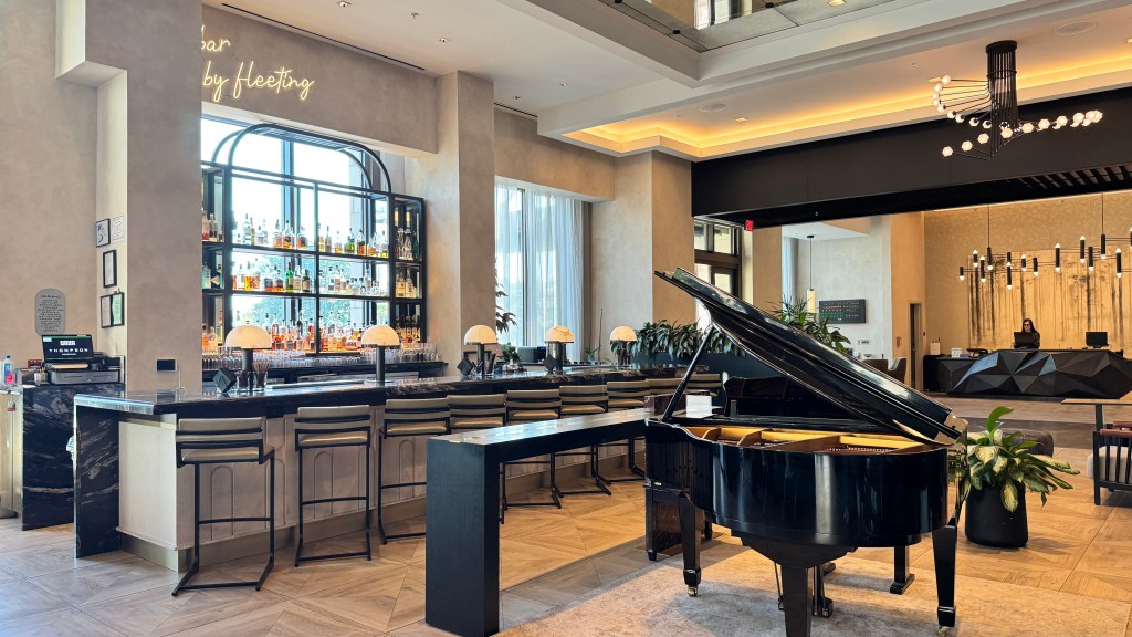Interior de um bar e lobby de hotel moderno, com um balcão de mármore escuro e banquetas claras à esquerda, uma estante de bebidas iluminada ao fundo e um piano de cauda preto à direita. No fundo, um balcão de recepção geométrico com uma pessoa sentada e lustres contemporâneos pendurados no teto alto