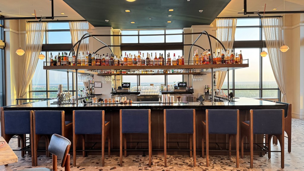 Bar moderno com balcão preto e oito banquetas de madeira com assentos azuis. Acima do balcão, uma prateleira suspensa exibe garrafas de bebidas. Ao fundo, janelas amplas com cortinas claras revelam uma paisagem verde e o céu claro. O teto é escuro, e o piso tem um padrão de mosaico