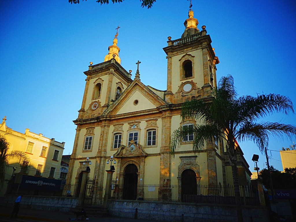 Igreja barroca de cor creme com duas torres sineiras e cruzes no topo, sob um céu azul intenso. Uma palmeira alta está à direita, e um prédio amarelo com a inscrição LIVRARIA SANTIAGO aparece à esquerda.